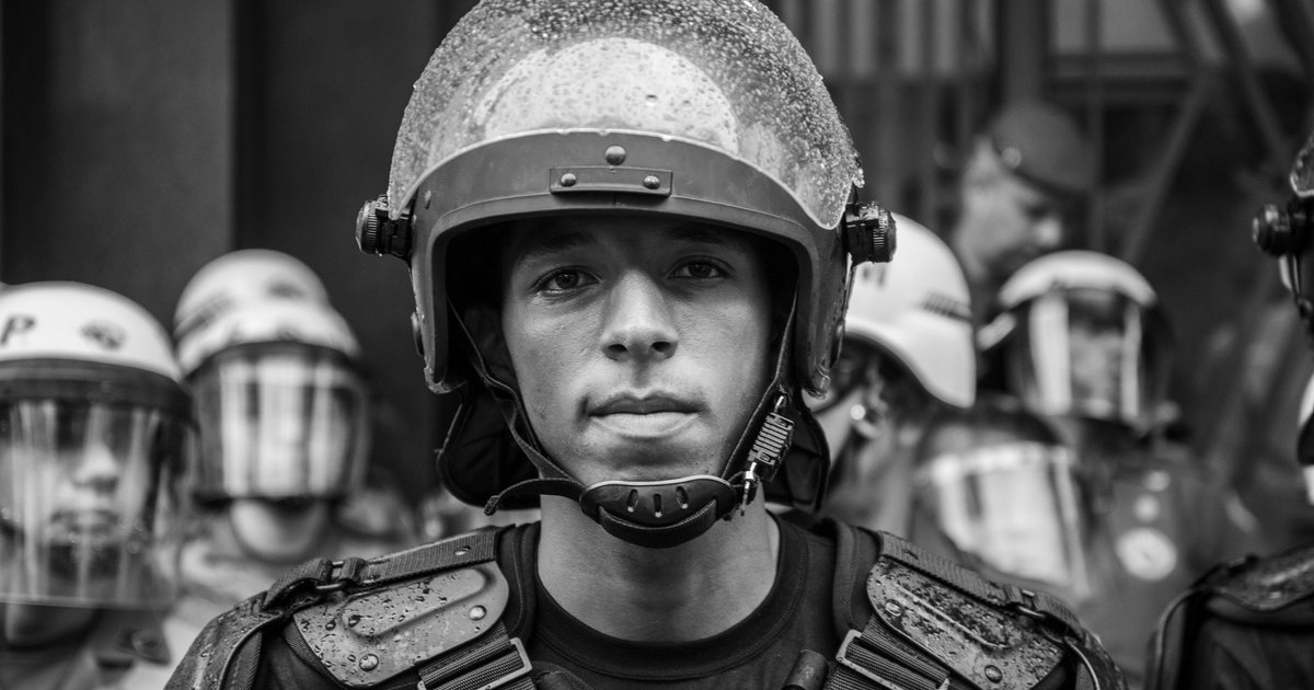 Riot police line facing a crowd of protesters on a street at dusk